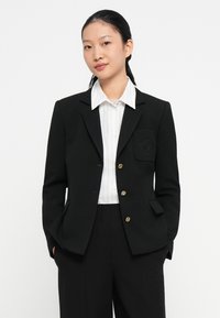 Blazer noir avec des boutons dorés, poche poitrine unique avec un motif subtil, associé à une chemise rayée blanche et un pantalon noir.
