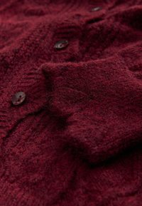 Bordeauxroter, gestrickter Pullover mit weichem, strukturiertem Stoff, sichtbaren Maschen und dunklen Knöpfen, flach liegend mit Falten.