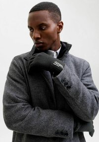 Abrigo gris de espiga con cuello de muesca, botones negros y detalle de cuello en capas, usado con un guante negro que presenta texto.