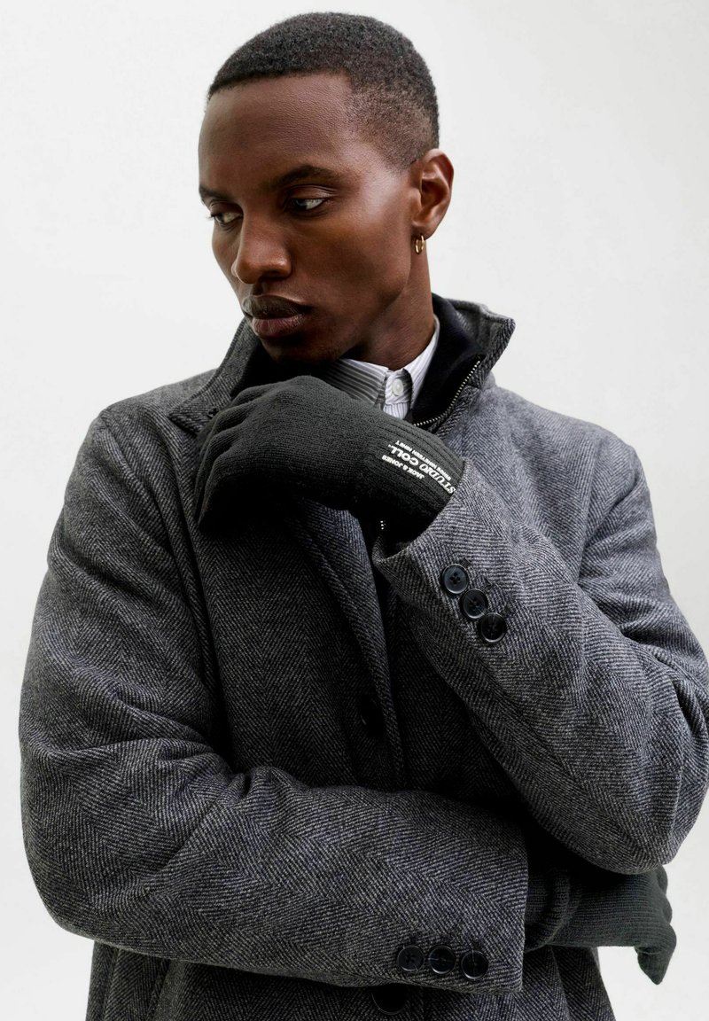 Abrigo gris de espiga con cuello de muesca, botones negros y detalle de cuello en capas, usado con un guante negro que presenta texto.