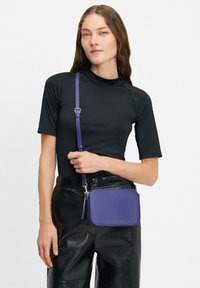 Borsa a tracolla in pelle viola con tracolla rimovibile, forma rettangolare, accessori in argento e texture liscia, abbinata a un top nero e pantaloni in pelle.