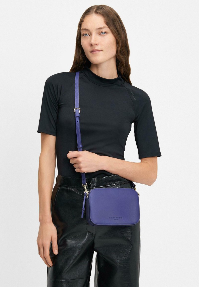 Borsa a tracolla in pelle viola con tracolla rimovibile, forma rettangolare, accessori in argento e texture liscia, abbinata a un top nero e pantaloni in pelle.