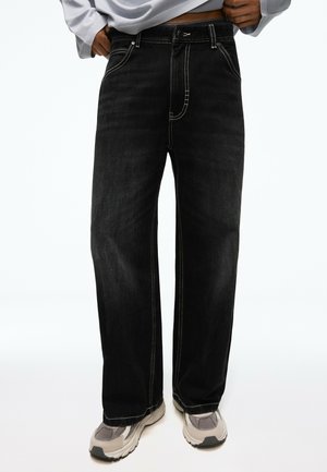 Personne portant un jean noir ample avec des surpiqûres blanches et des baskets gris clair, debout devant un fond blanc.