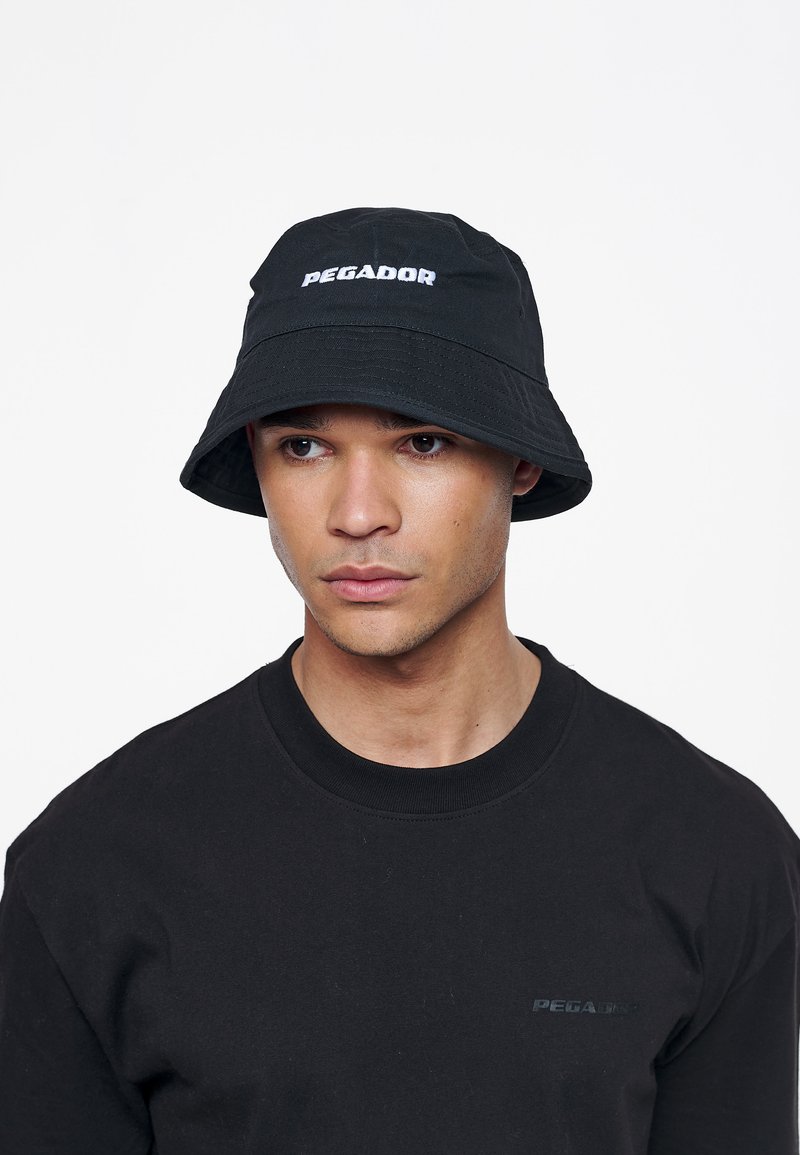 Fekete vödörkalap, fehér "PEGADOR" logóval, sima textúrájú anyagból. Fekete kerek nyakú pólóval viselve.