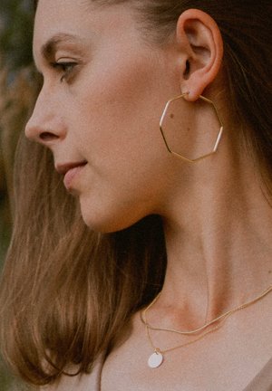 Goldene oktagonale Creolen und geschichtete goldene Halsketten. Die Ohrringe haben ein dünnes, geometrisches Design. Das Modell hat braunes Haar und einen glatten Hals.