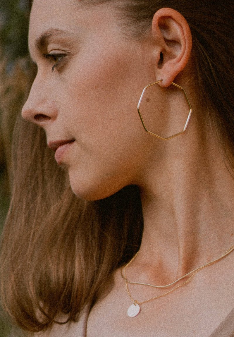 Goldene oktagonale Creolen und geschichtete goldene Halsketten. Die Ohrringe haben ein dünnes, geometrisches Design. Das Modell hat braunes Haar und einen glatten Hals.