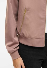 Prim-plan cu mâneca unei jachete mov și buzunar cu fermoar auriu, purtată peste pantaloni negri, mâna relaxată pe lângă corp.