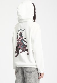 Haut à capuche blanc surdimensionné avec un imprimé graphique représentant une figure et un tigre au dos. Manches longues, texture douce et capuche avec cordon de serrage.