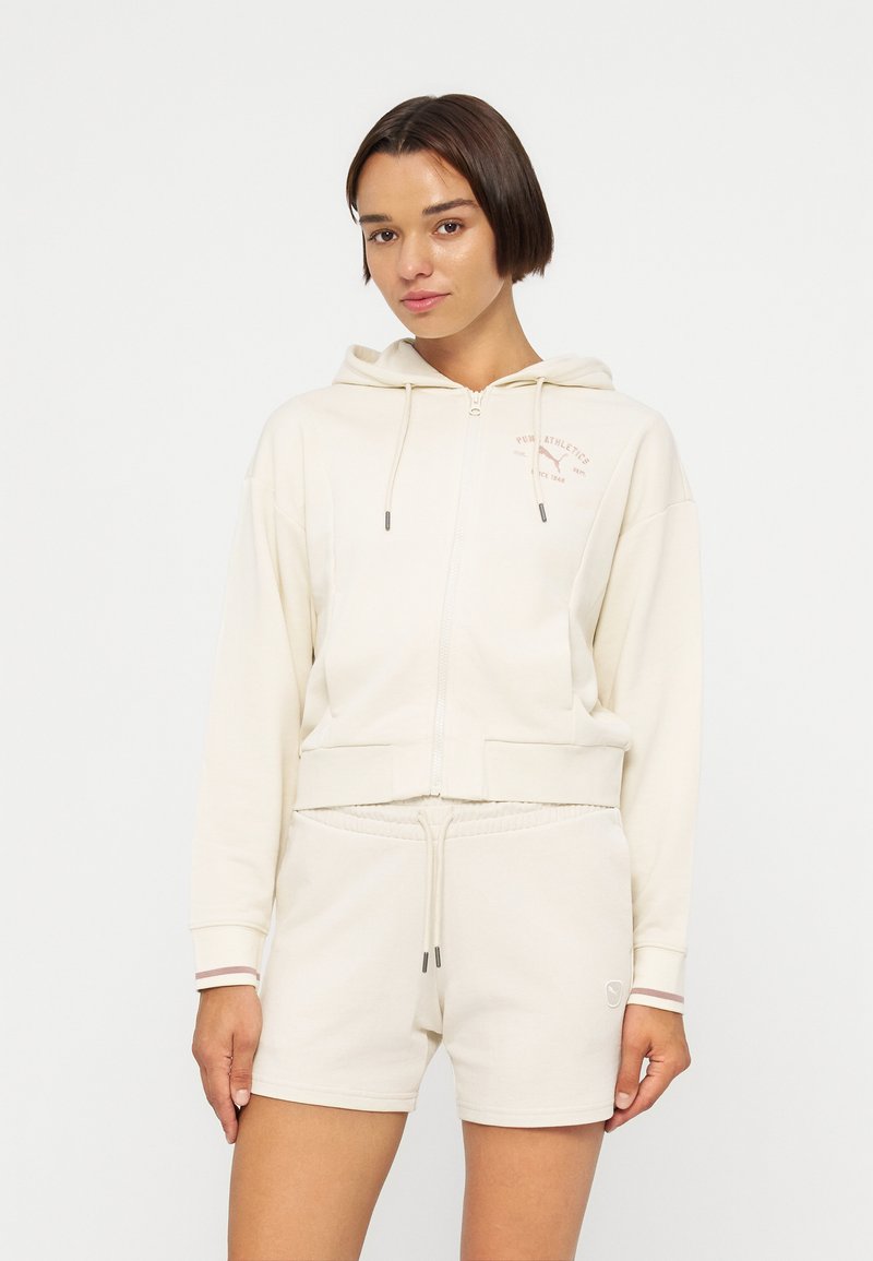 Femme portant un sweat à capuche crème à fermeture éclair avec cordons et un short crème assorti, debout devant un fond blanc uni.