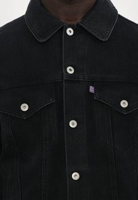 Veste en denim noir avec un col pointu, boutons-pression en argent et deux poches poitrine avec un petit logo rayé en accent.