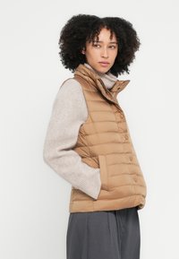 Bēšīga pufjakas veste ar augstu apkakli, pogu aizdari un divām sānu kabatām. Mīksta, kviltēta tekstūra, uzvilkta pār gaiši bēšu džemperi.