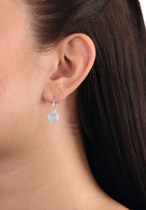 Orecchio sinistro di una donna con capelli castano scuro, che indossa un piccolo orecchino a cerchio in argento con un pendente rotondo e testurizzato.