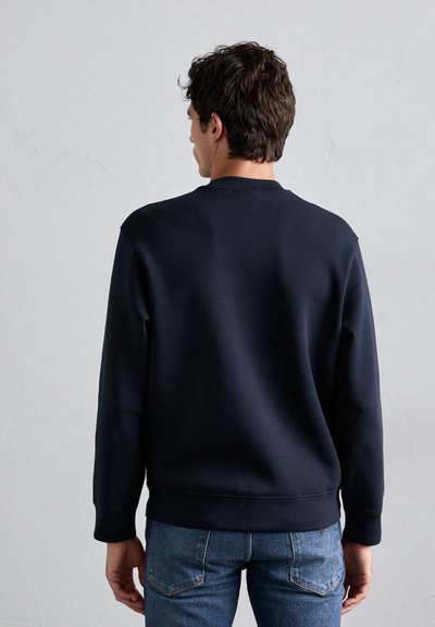Man in een eenvoudige donkerblauwe sweatshirt en blauwe spijkerbroek, staande met zijn rug naar een lichtgrijze muur.