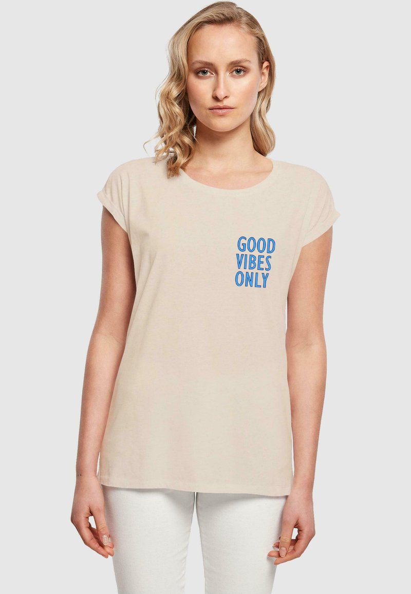 Beige Baumwoll-T-Shirt mit kurzen umgeschlagenen Ärmeln. Auf der Vorderseite befindet sich blauer Text "NUR GUTE VIBES". Leichter, lockerer Schnitt, glatte Textur.
