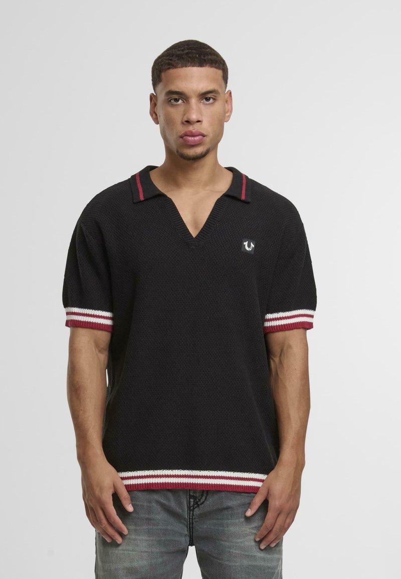 Polo de manga corta negro con escote en V, puños a rayas rojas y blancas, y un detalle de logo en el lado izquierdo del pecho. Acabado de tejido texturizado.