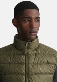 Chaleco acolchado de color verde oliva con alto collar y cremallera frontal. Presenta un logo de marca en el pecho y tela gris contrastante en los laterales.