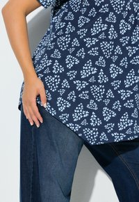 Granatowa bluzka z białym, kwiatowym wzorem w kształcie serca, zestawiona z ciemnoniebieskimi jeansami. Skupienie na fakturze tkaniny i warstwowej sylwetce.