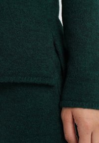 Gros plan d'une main reposant à côté d'une manche de manteau en laine vert foncé avec une couture de poche visible.