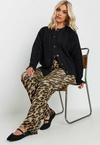 Cardigan nero lavorato a maglia con design a trecce, abbinato a pantaloni marroni con stampa leopardata. Scarpe nere senza tacco completano il look.