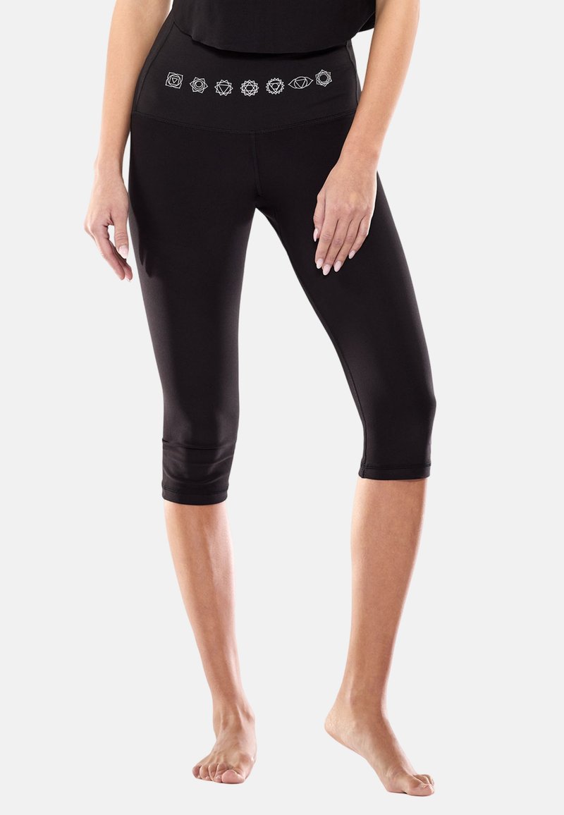 Kvinde iført sorte kropsnære leggings med hvide chakra-symboler på taljen, står barfodet på en lys baggrund.