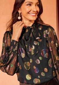 Blusa nera trasparente con motivi rotondi iridescenti in oro, verde e borgogna. Presenta un collo a fiocco e maniche arricciate.