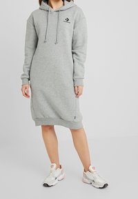 Vestido sudadera gris hecho de material suave, con un bolsillo tipo canguro, capucha con cordón y un acento de logo, combinado con zapatillas blancas.