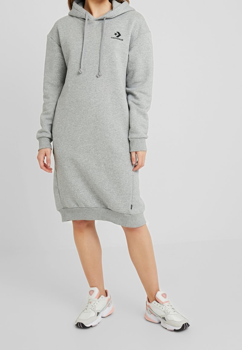 Vestido sudadera gris hecho de material suave, con un bolsillo tipo canguro, capucha con cordón y un acento de logo, combinado con zapatillas blancas.