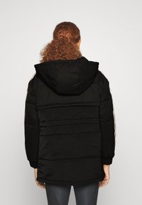 Giacca nera imbottita con cappuccio, dotata di polsini elasticizzati e strisce decorative sui lati. Il tessuto ha una finitura opaca e una texture trapuntata.