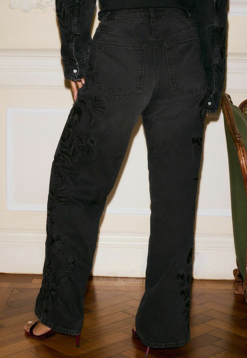 Personne portant un jean noir taille haute avec broderies florales, associé à une veste noire brodée et des chaussures à talons hauts rouge foncé.