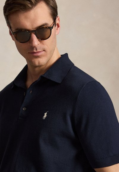 Άνδρας που φοράει γυαλιά ηλίου από χελώνα και μπλε πόλο πουκάμισο με μικρό λευκό κεντητό λογότυπο αλόγου.