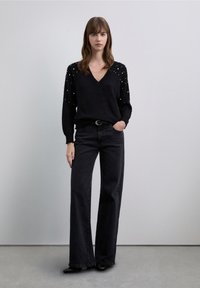 Svart stickad tröja med V-ringning och pärldetaljer på �ärmarna, kombinerad med svart vida jeans med hög midja och svarta skor.