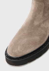 Bottine en suede beige à bout arrondi, avec une semelle en caoutchouc noir contrastante et des bords ondulés, et des détails de couture minimalistes.