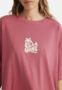 Rose-colored, oversized cotton t-shirt featuring a textured white logo with palm tree design, positioned centrally on the chest area.