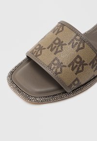 Παντόφλα με ανοιχτή μύτη, με καφέ ιμάντα από ύφασμα μονογράμματος και περιμετρικό διακοσμητικό με στρας γύρω από την τετράγωνη άκρη της σόλας.