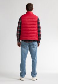 Rote Steppweste mit horizontalem Karomuster, getragen über einem karierten Hemd, kombiniert mit hellblauen Jeans und weißen Turnschuhen.