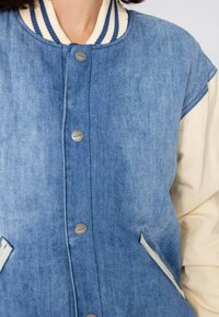 Veste varsity en denim avec un corps bleu clair, des manches crème, un col côtelé bleu marine et crème, des boutons-pression en argent et des poches latérales.
