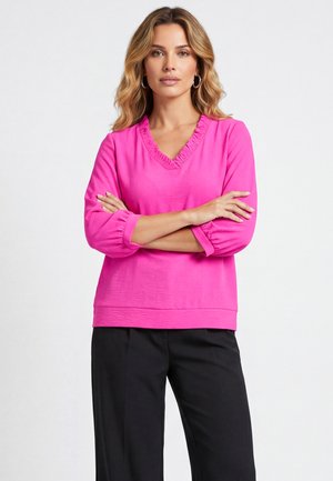 Kobieta o falowanych blond włosach, ubrana w jasnoróżową bluzkę z dekoltem w serek z falbanką i czarne spodnie, ze skrzyżowanymi rękami, stojąca na jednolitym tle.