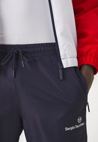 Navy track pants with elastic waistband and drawstring. Material features a subtle texture. Logo displayed on the left side.