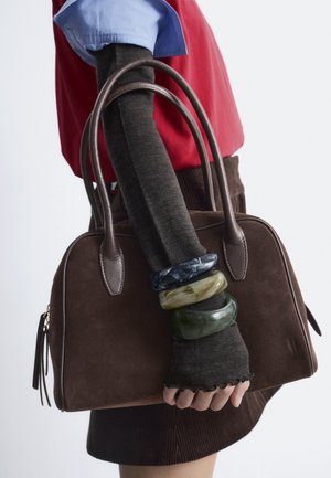 Brown suede handbag with rounded handles, paired with a dark, textured sweater and colorful bangles on a slender arm, against a light background.
