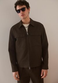 Chaqueta marrón con cremallera y dos bolsillos en el pecho, hecha de una tela texturizada. Llevada sobre una camisa clara con una cadena de plata.