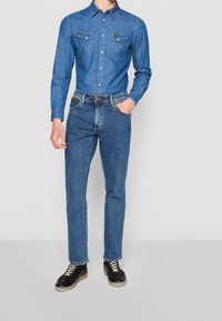 Camisa de mezclilla con cuello abotonado, dos bolsillos en el pecho, vaqueros azules y zapatos negros. Presenta costuras en contraste y un corte recto.