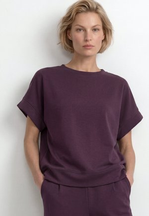 Blonde vrouw met kort haar, gekleed in een losse donkerpaarse top met korte mouwen en bijpassende broek, staand met handen in de zakken tegen een witte muur.