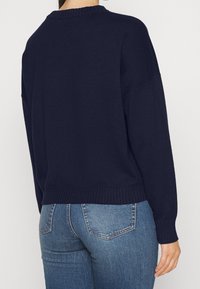 Maglione lavorato a maglia blu navy con collo e orlo a coste. Presenta spalle abbassate e una vestibilità rilassata. Abbinato a jeans blu chiaro.