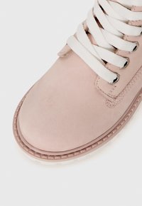 Primo piano di una punta di stivale scamosciato rosa chiaro con lacci bianchi infilati attraverso occhielli metallici su uno sfondo bianco.