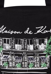 Sac fourre-tout noir présentant un croquis blanc d'une vitrine avec des plantes vertes et des auvents, étiqueté "Maison de Karl". Tissu et bandoulière solides.