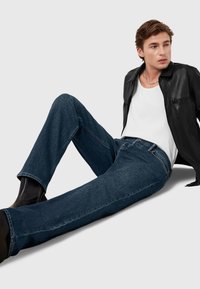 Vaqueros de denim azul con corte recto, combinados con una camiseta sin mangas blanca y una chaqueta de cuero negra. La textura es suave y ligeramente desgastada.