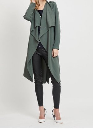 Femme portant un long manteau drapé vert foncé à devant ouvert, un haut et un pantalon noirs, ainsi que des chaussures blanches à talons hauts, debout devant un fond uni.