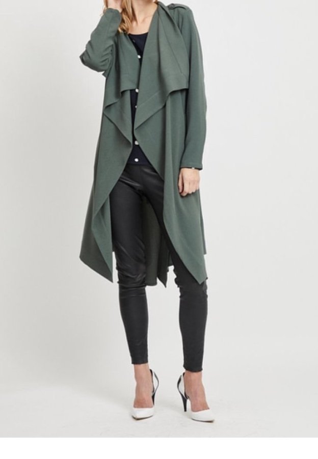 Femme portant un long manteau drapé vert foncé à devant ouvert, un haut et un pantalon noirs, ainsi que des chaussures blanches à talons hauts, debout devant un fond uni.