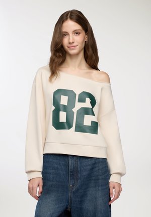 Felpa color crema con spalle scoperte, caratterizzata da una grande stampa verde "82", polsini a coste e un design cropped, abbinata a jeans di denim blu scuro.