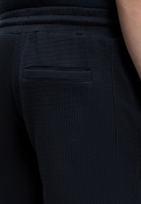 Gros plan d’un pantalon texturé foncé avec une poche passepoilée à l’arrière et une partie du bras d’une personne visible sur le côté droit.